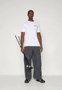 Bela kratka majica s kratkimi rokavi z logotipom "Terrex", ohlapne sivo cargo hlače z rumenim šivom, črna obutev, drži dva pohodniška palica.