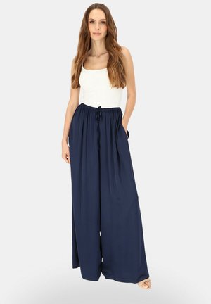 Kobieta o długich brązowych włosach, ubrana w biały top na ramiączkach oraz luźne granatowe spodnie z szerokimi nogawkami i sznurkiem w pasie.