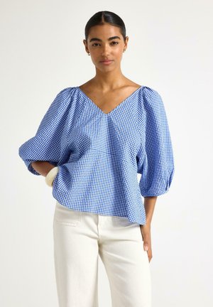 Donna che indossa una camicetta a quadri blu e bianca con scollo a V e maniche a sbuffo e pantaloni bianchi, in piedi davanti a uno sfondo semplice.