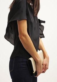 Sort kortærmet top med en svævende sort chiffon-accent, parret med en tofarvet clutch - creme og sort - som holdes i hænderne over mørke jeans.