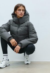 Gray puffer jacket with a high collar and zip front, featuring horizontal stitching and a soft, smooth texture. Worn with black leggings and sneakers.