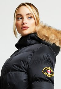 Svart dunjakke med hette kantet med pels, med en gul og rød brodert logo på venstre erme og glidelås foran.