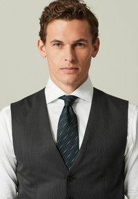 Graue Weste mit Revers und zwei Knöpfen, getragen über einem weißen Hemd. Eine dunkelblau-grüne Krawatte mit subtilen Streifen rundet das Outfit ab.