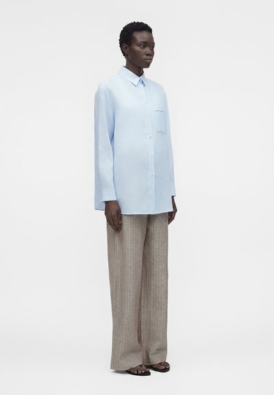 Άτομο που φοράει oversized πουκάμισο ανοιχτού μπλε χρώματος με κουμπιά και μπεζ παντελόνι με φαρδιά μπατζάκια και ρίγες, στέκεται μπροστά σε λευκό φόντο.