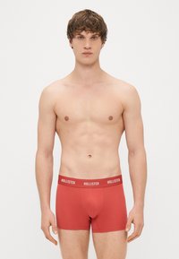 Boxers de algodão vermelhos com um ajuste justo, apresentando uma cinta elástica branca com a marca mostrando "HOLLISTER". Textura suave, comprimento até a coxa.