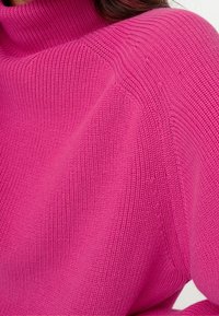 Pull en tricot rose vif avec un col haut côtelé. Présente une surface texturée avec une coupe ample et des manches larges.