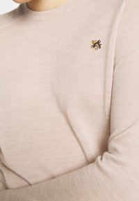 Un pull rose clair en tissu doux, avec un col rond et un petit détail de broderie florale sur le côté gauche de la poitrine.