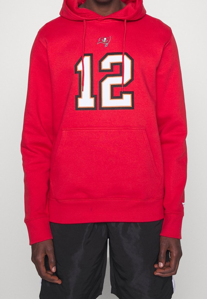 Personne portant un sweat à capuche rouge avec le numéro 12 et un petit logo de drapeau pirate au-dessus, assorti à un short noir.