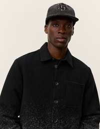 Junger schwarzer Mann mit einer dunkelgrauen gemusterten Mütze und einer schwarzen, geknöpften Jacke mit einer Brusttasche und einem gesprenkelten Farbverlauf.