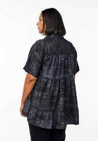 Woman with straight black hair wearing a loose, short-sleeve, dark patterned tunic facing left on a white background.