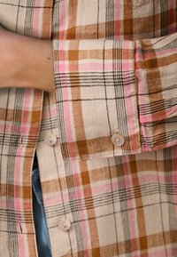 Košile na knoflíky s kostkovaným vzorem v béžové, růžové a hnědé barvě; bavlněná látka s jemnou texturou; má kulaté knoflíky a detail na manžetách.
