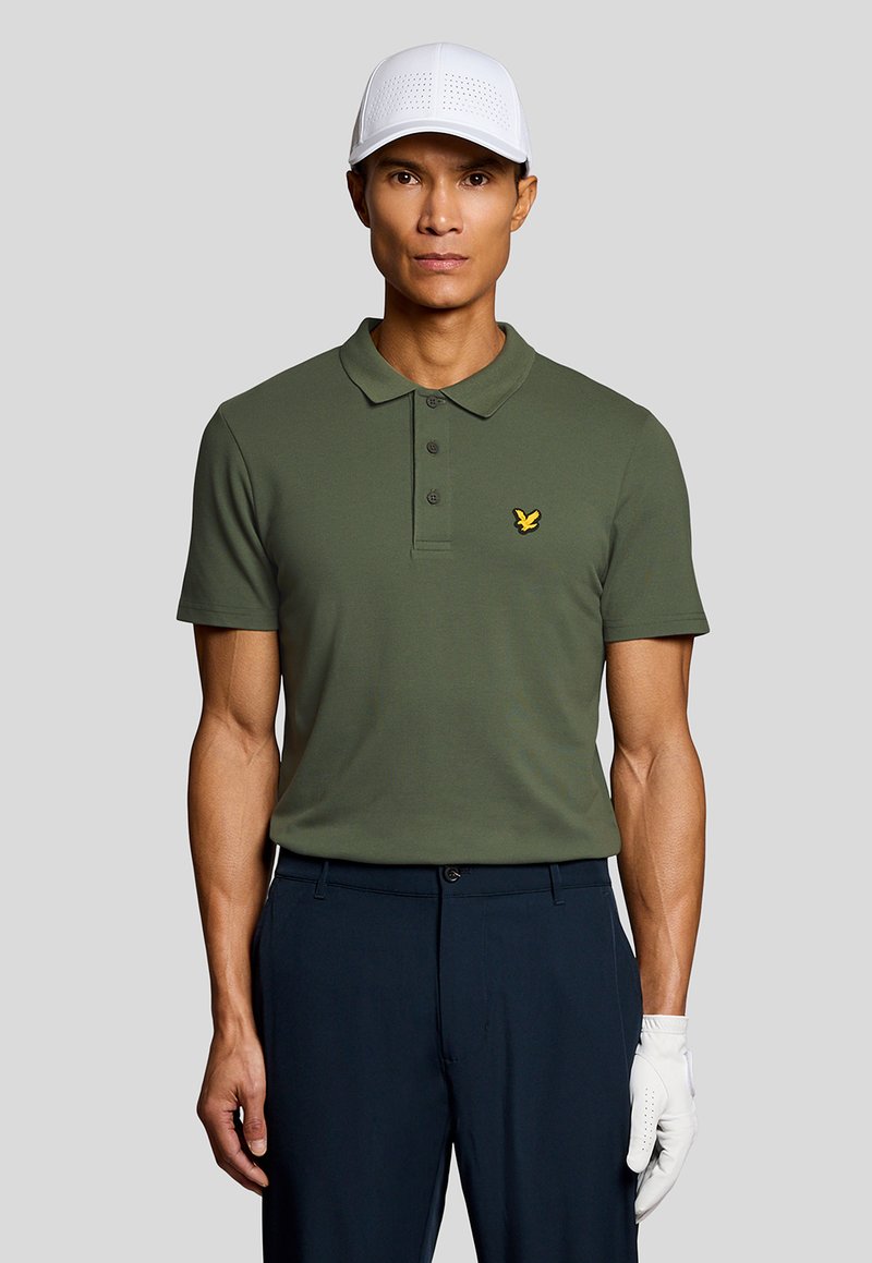 Mann mit weißer Kappe, olivgrünem Poloshirt mit gelbem Vogel-Logo, dunkelblauer Hose und weißem Golfhandschuh vor einfarbigem Hintergrund.