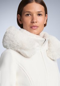 Jachetă albă cu un finisaj texturat, având un guler pufos din blană albă. Se observă închiderea cu fermoar și detalii de design elegante.