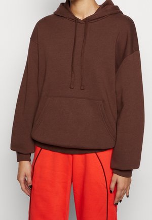 Person iført en brun hættetrøje med snoretræk og en kængurulomme, kombineret med lyse orange bukser med sorte kanter.