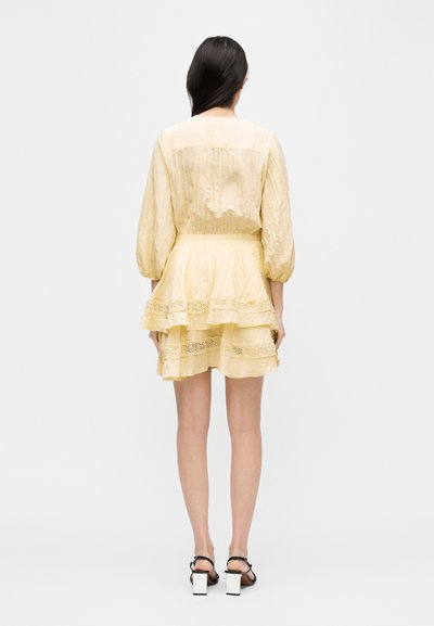 Γυναίκα με μακριά σκούρα μαλλιά, που φοράει ένα ανοιχτό κίτρινο φόρεμα με βολάν και λεπτομέρειες από δαντέλα, καθώς και φούστες με φουσκωτά μανίκια, συνδυασμένο με μαύρα και λευκά σανδάλια με χοντρά τακούνια.