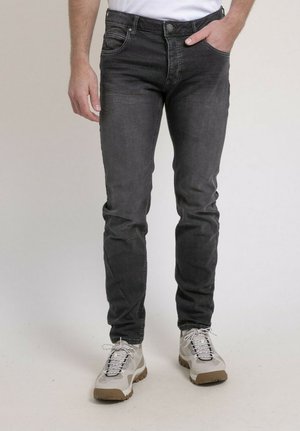 Homme portant un jean noir ajusté et des baskets blanches, debout avec une main dans la poche devant un fond uni.