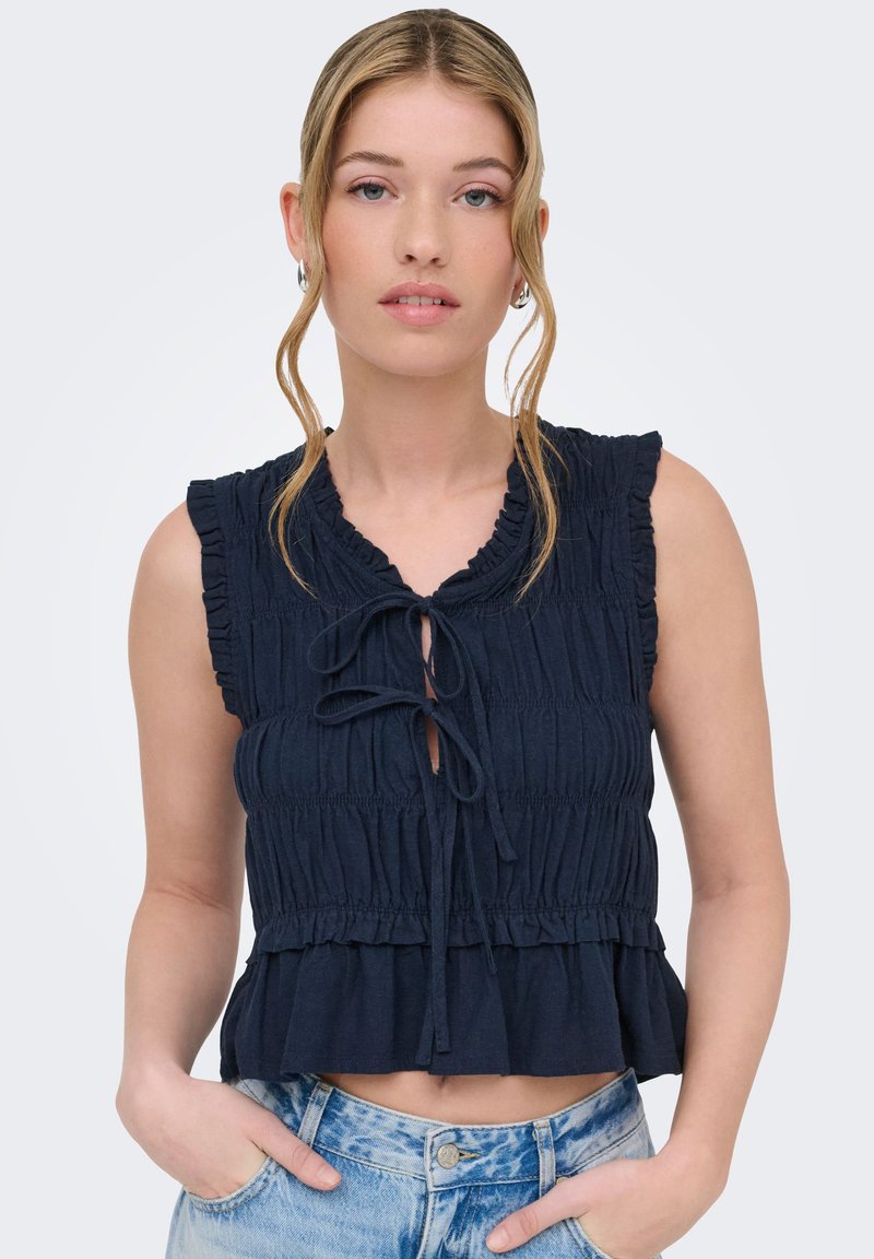 Jeune femme aux cheveux blonds porte un haut sans manches bleu marine froncé avec des lacets à l'avant et un jean en denim bleu clair, les mains dans les poches.