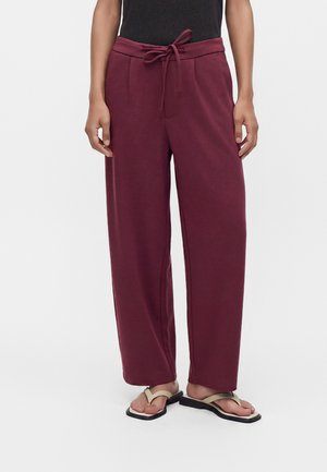 Person wearing wide-leg maroon pants with a tied waist and beige sandals with black soles standing against a plain background.