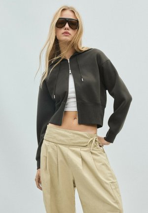 Femme blonde portant de grandes lunettes de soleil noires, un sweat à capuche noir court zippé, un crop top blanc et un pantalon ample beige taille haute sur un fond uni.
