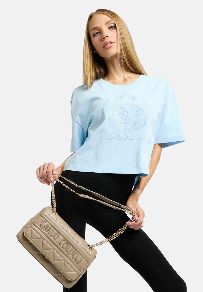Lyseblå kort t-shirt med præget logodesign, kombineret med en beige quiltet håndtaske med kæderem og logo, båret med sorte leggings.