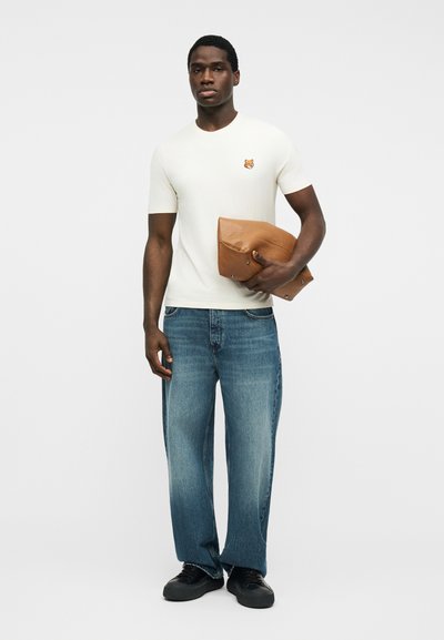 Άνδρας που φοράει λευκό πουκάμισο με κοντά μανίκια και λογότυπο τίγρης, μπλε τζιν και μαύρα παπούτσια, κρατώντας μια διπλωμένη καφέ δερμάτινη τσάντα.