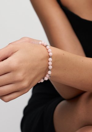 Nahaufnahme eines Handgelenks mit einem Armband aus pinken und silbernen Perlen, vor einem Hintergrund aus verschränkten Armen und schwarzer Kleidung.