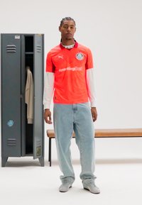 Rdeč dres z belo majico z dolgimi rokavi pod njim, z logotipom in besedilom. Svetlo modre ohlapne kavbojke in sive superge, stoji ob omarici.