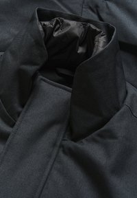 Tumšs, teksturēts auduma apkakle ar spīdīgu melnu oderi, demonstrējot modernu dizainu un strukturētu formu tuvplānā.