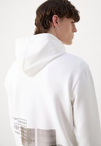 Sweat à capuche blanc avec une capuche texturée et bordée de blanc et un détail graphique présentant des motifs géométriques dans des tons doux au dos.