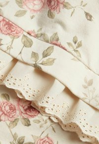 Kangas kuviolla, jossa on vaaleanpunaisia ruusuja beigellä taustalla. Sisältää pitsireunuksen, jossa on rei'itettyjä kuvioita. Pehmeä, puuvillainen rakenne.