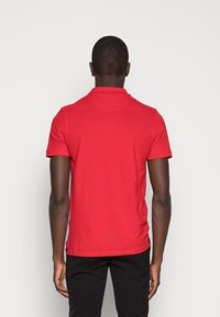 Punainen polo-paita, lyhyet hihat, ribatut kaulukset, normaali istuvuus, sileä puuvillakangas, sivuaukot helmassa liikkuvuuden helpottamiseksi.