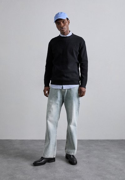 Zwarte gebreide trui, blauwe pet, gestreept coll shirt eronder, lichtgewassen baggy jeans, zwarte schoenen, staand tegen een neutrale achtergrond.