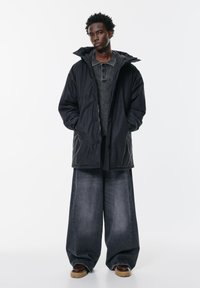 Tânăr îmbrăcat într-o jachetă neagră cu glugă, pulover gri, blugi foarte largi de culoare gri închis și pantofi maro, pe un fundal alb simplu.