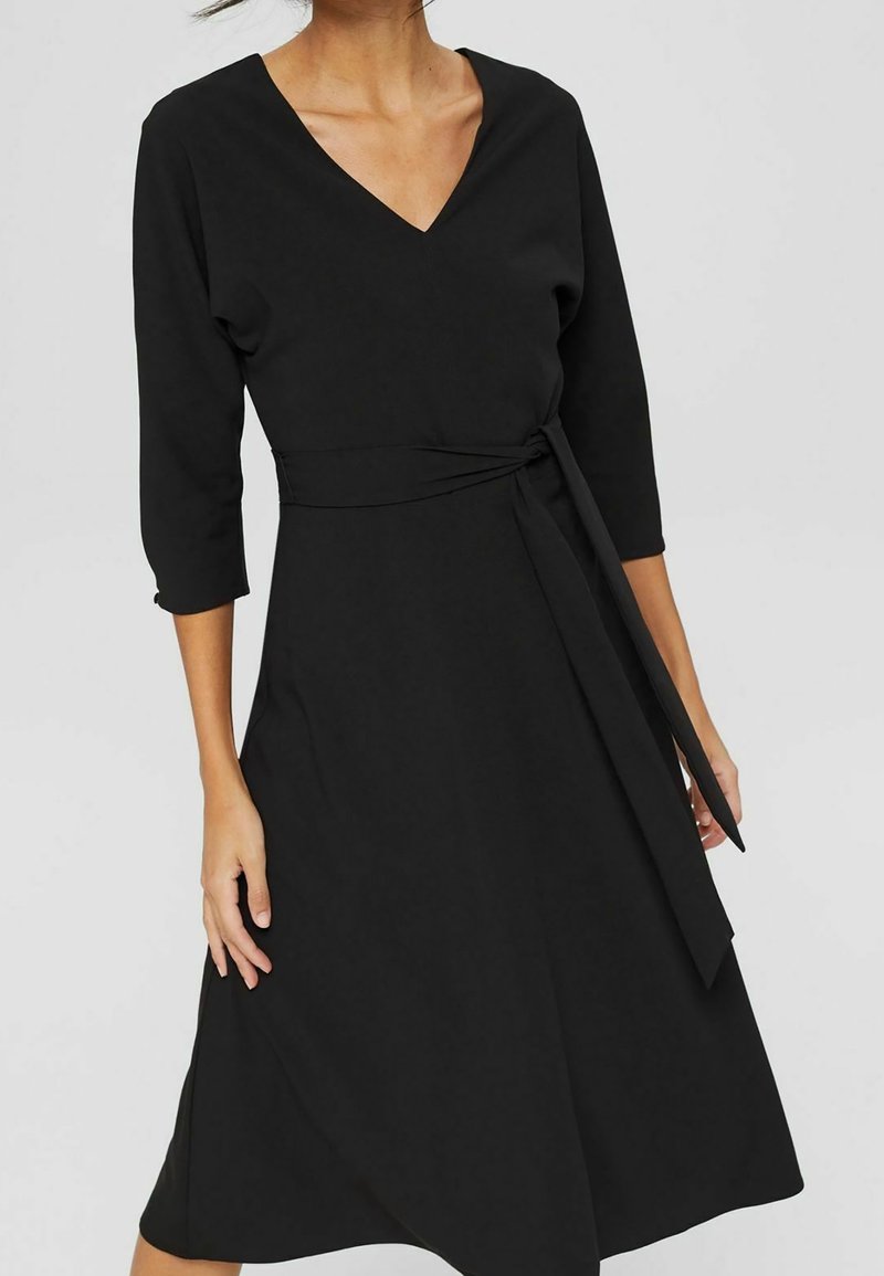 Femme portant une robe noire à col en V avec des manches trois-quarts et une ceinture nouée à la taille, debout devant un fond clair uni.