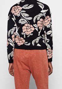 Schwarzer Pullover mit creme- und rosa floralem Muster, strukturiertem Strick und geripptem Saum. Kombiniert mit weichen, rostfarbenen Hosen mit glatter Oberfläche.