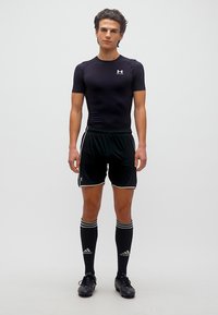 Νεαρός άνδρας που στέκεται με μαύρη αθλητική ενδυμασία με κοντά μανίκια, σόρτς, κάλτσες μέχρι το γόνατο και ποδοσφαιρικά παπούτσια σε λευκό φόντο.