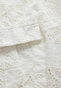 Tissu blanc avec une broderie florale découpée complexe et un bouton visible près d'un bord ou d'un poignet plié.