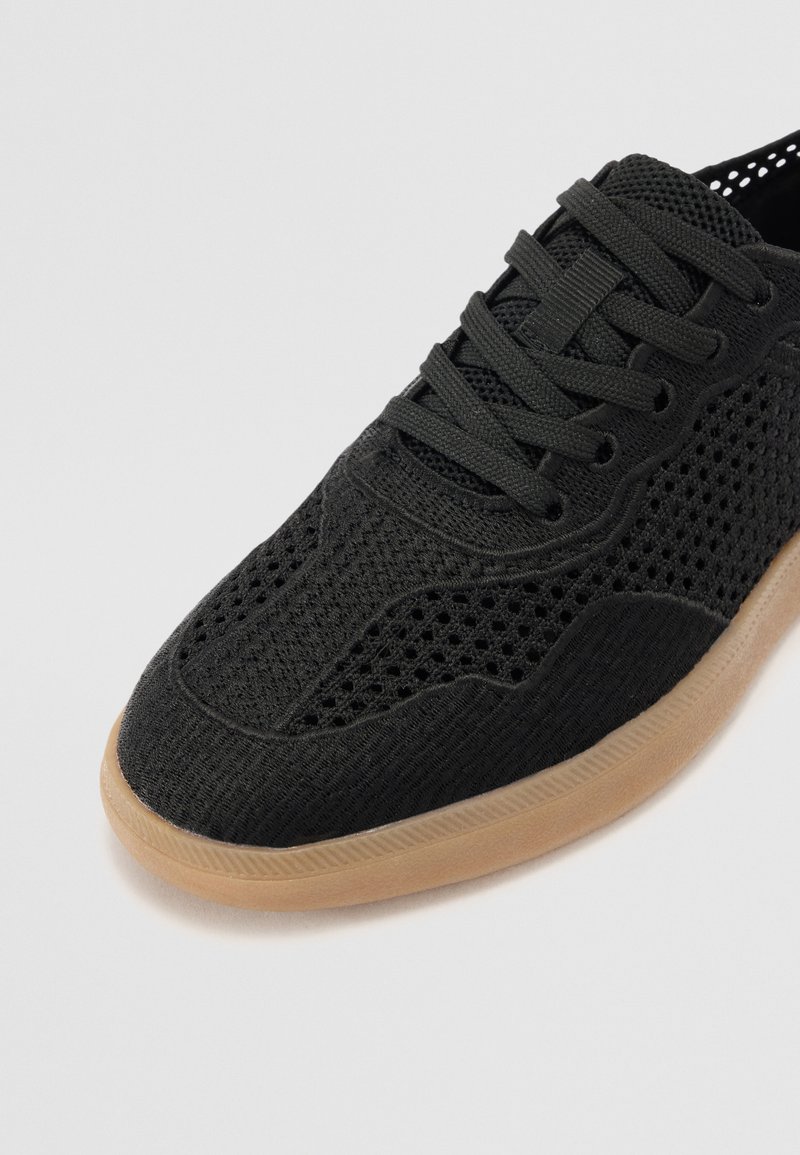 Zapatilla deportiva negra con parte superior de malla transpirable, punta redondeada, cierre de cordones y suela de goma con textura.