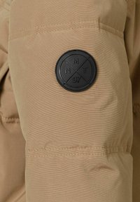 Tan puffjacka med ärmar som har en cirkulär svart tygpatch med pr�äglade mönster, inklusive "NMT" och "97." Slät textur med kontrasterande sömmar.