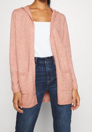 Kobieta ubrana w różowy kardigan z kapturem i otwartym przodem, nałożony na biały top oraz ciemnoniebieskie jeansy z wysokim stanem, stojąca na tle jednolitego tła.