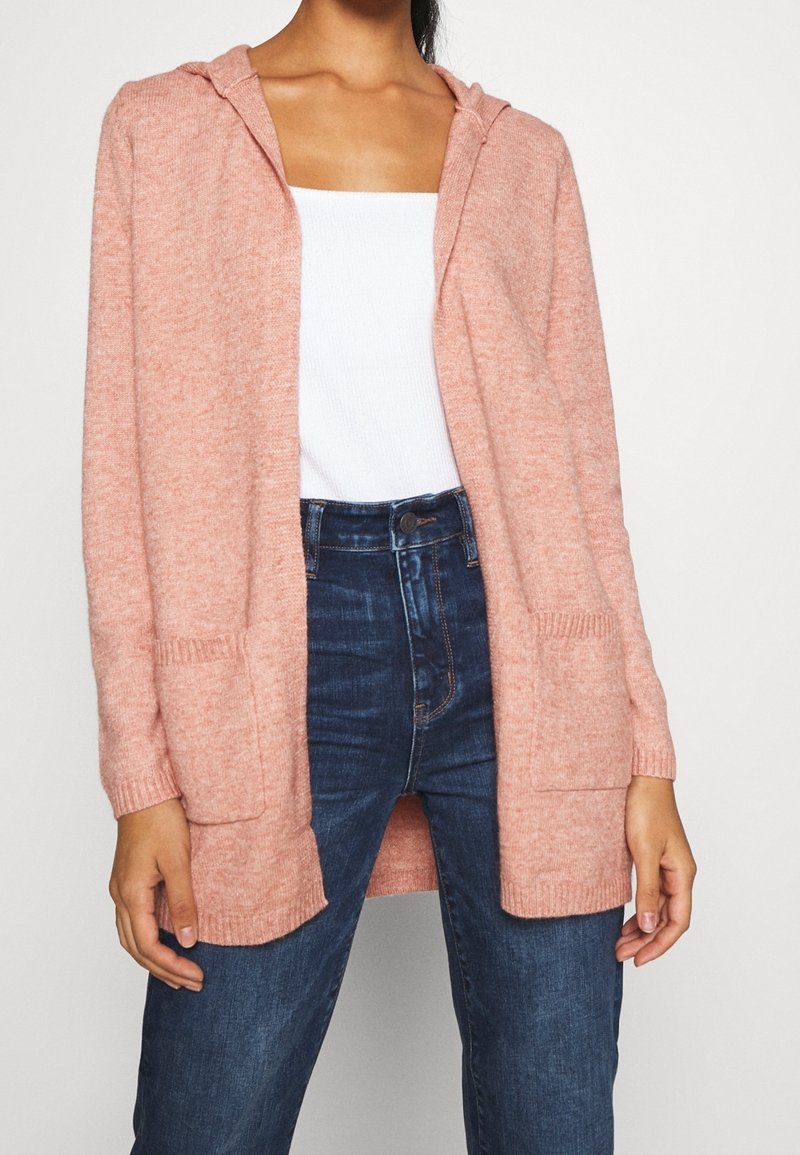 Femme portant un cardigan rose à capuche ouvert sur un haut blanc et un jean bleu foncé taille haute, debout devant un fond uni.