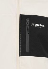 Gros plan sur une veste avec une poche à fermeture éclair noire et un logo blanc "JJ Studios Jack & Jones" sur un fond de tissu beige.