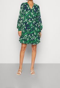Kwiatowa sukienka o kroju kopertowym z długimi rękawami, głębokim dekoltem w kształcie litery V i falbaniastym dołem. Zielone liście na granatowym tle. Połączona z białymi sandałami na cienkich obcasach.