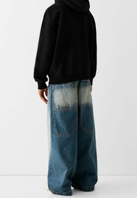 Persona con pantalones de mezclilla azules de dos tonos y holgados, y una sudadera negra, de pie contra un fondo blanco liso.