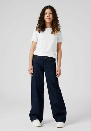 Dunkelblaue Jeans mit weitem Bein und kontrastierenden Nähten, kombiniert mit einem taillierten weißen Baumwoll-T-Shirt und weißen Turnschuhen, auf einem neutralen Hintergrund präsentiert.