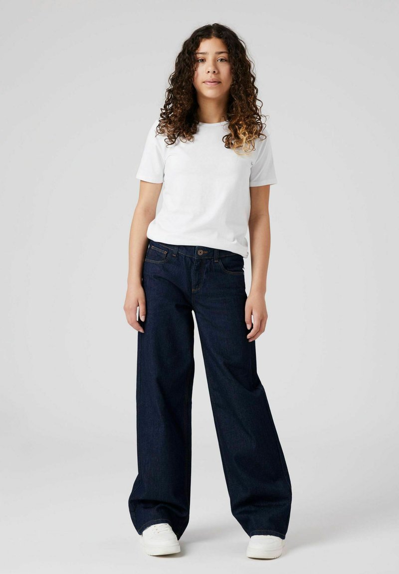 Tummansiniset leveälahkeiset farkut kontrastitikkauksilla, yhdistettynä istuvaan valkoiseen puuvillapaitaan ja valkoisiin lenkkareihin, esitetty neutraalilla taustalla.