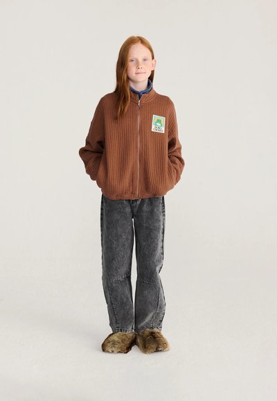 Jeune personne aux longs cheveux roux portant une veste marron zippée, un jean gris et des chaussons marron fourrés, debout les mains dans les poches.