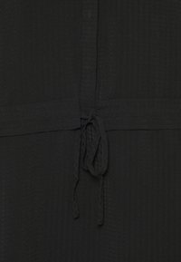 Vêtement noir en tissu texturé. Doté d'une fermeture à nouer à la taille, avec des coutures visibles et un motif vertical subtil.