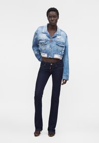 Femme portant une veste en jean bleu clair courte et usée ainsi qu'un jean évasé bleu foncé, debout avec une main le long du corps.