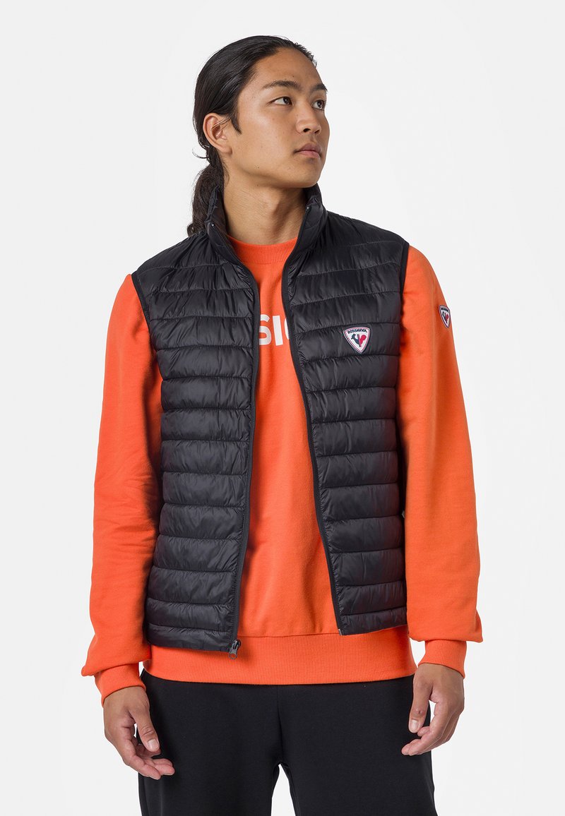 Černá vyp padded vesta s vertikálním prošíváním, vysokým límcem, bočními kapsami a logem na levé hrudi, nošená přes oranžovou mikinu.